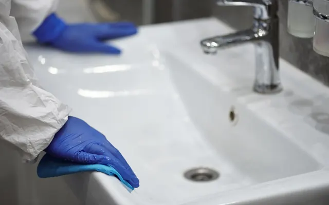 Gloved hands deep-cleaning a white bathroom sink and chrome faucet