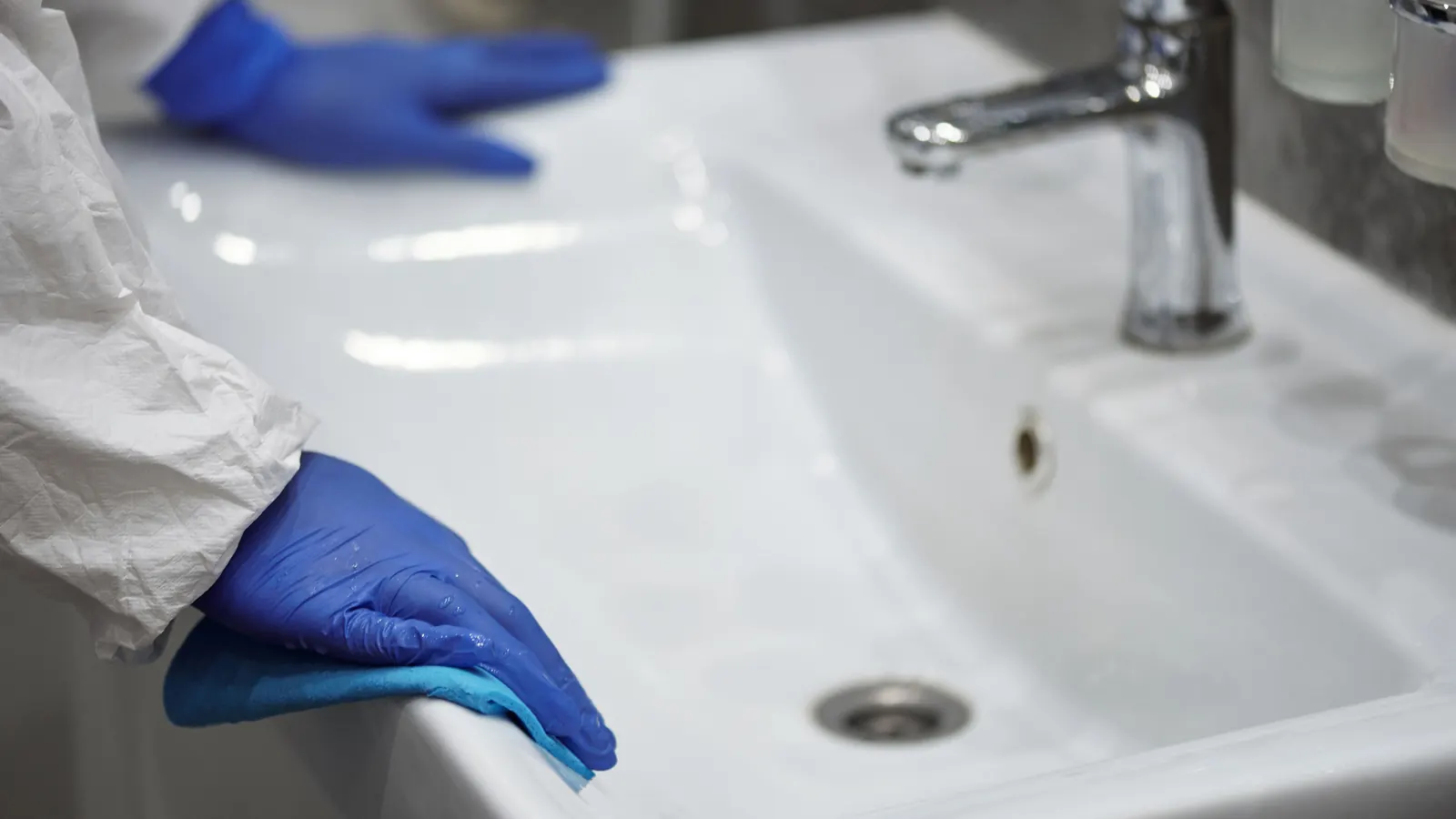 Gloved hands deep-cleaning a white bathroom sink and chrome faucet