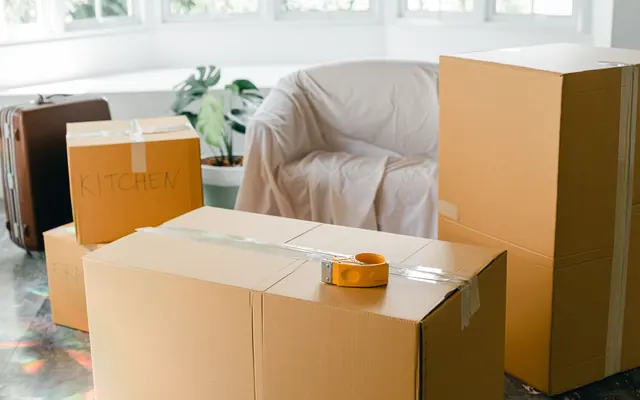 Taped moving boxes beside a sheet-covered armchair in a sunlit apartment on move-out day