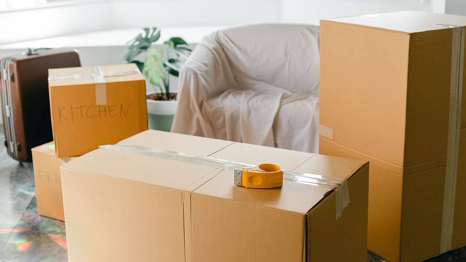 Taped moving boxes beside a sheet-covered armchair in a sunlit apartment on move-out day