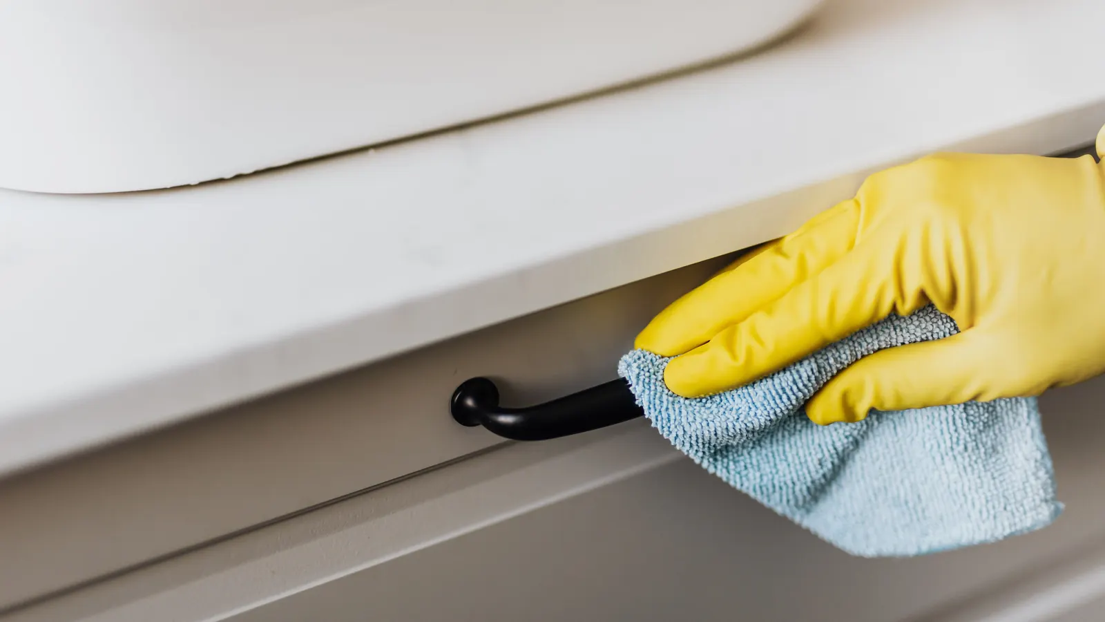 Gloved hand wiping a cabinet drawer front and handle with a microfiber cloth