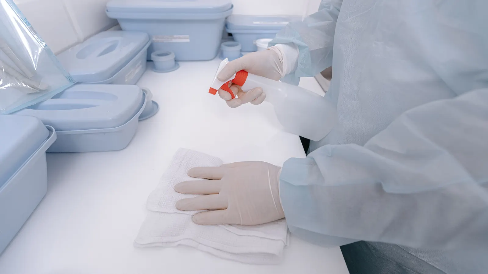 Gloved hands disinfecting a clinic counter with spray and a cloth beside sterile trays