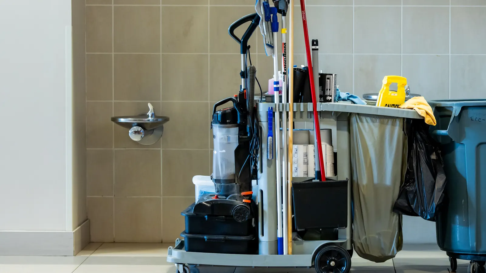 Stocked janitorial cart with mops, vacuum and trash bin in a tiled commercial corridor