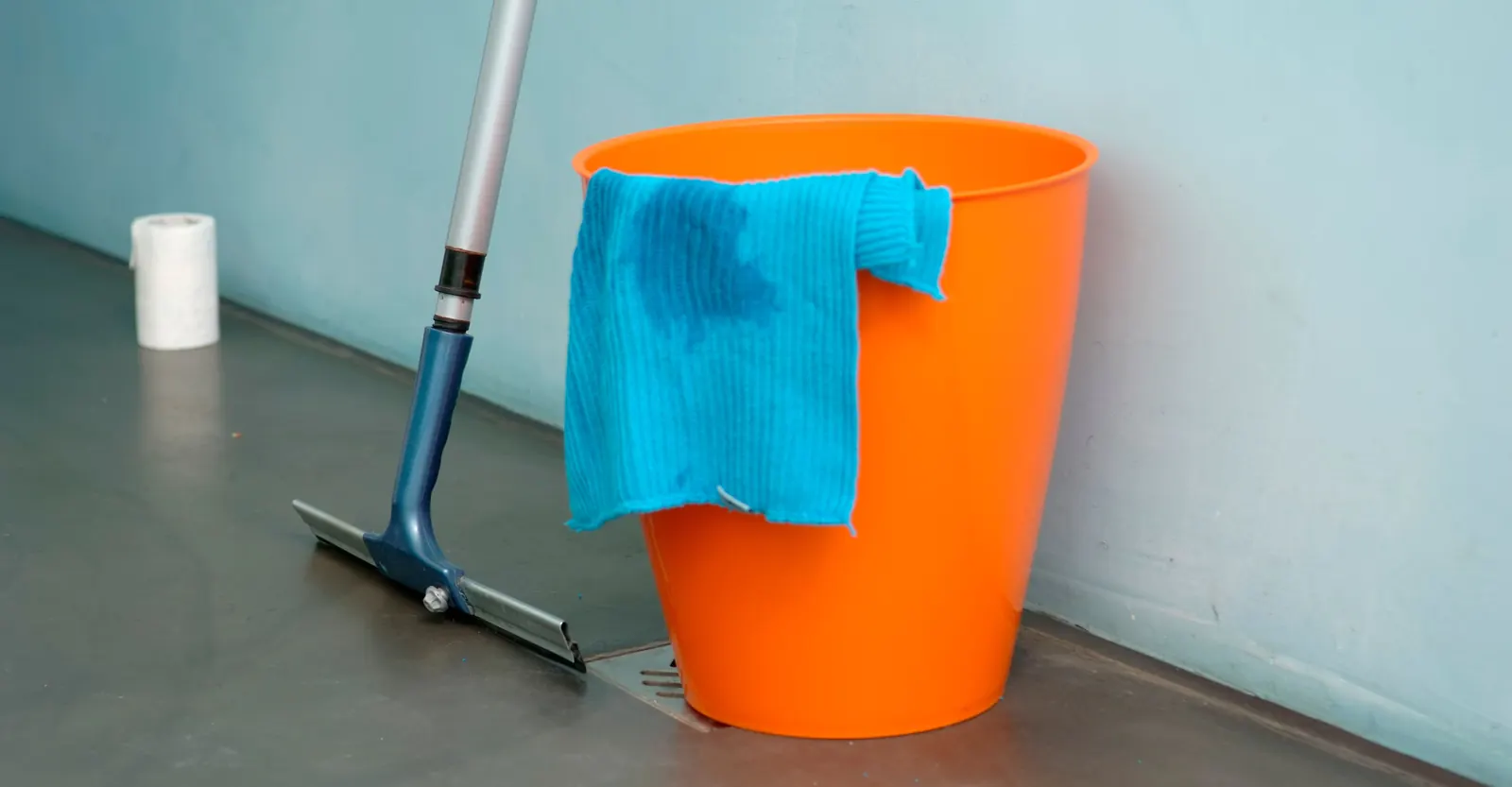 Orange bucket with a blue cleaning cloth and a floor squeegee on a polished concrete floor